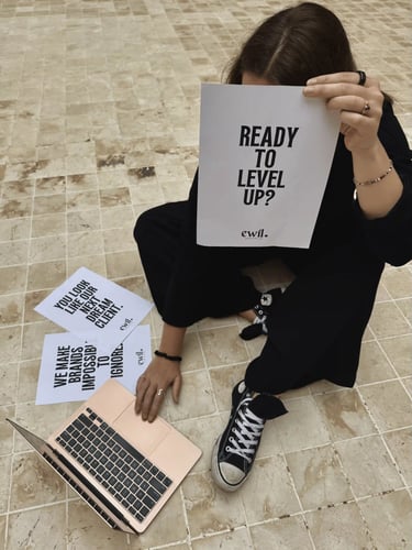 A creative professional holds a sign saying Ready To Level Up next to a laptop and marketing flyers.