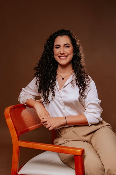 Smiling woman with long curly hair wearing a white silk blouse and tan trousers sitting in a chair.
