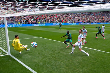 A soccer player kicks a ball into the goal past a goalkeeper during a match in a crowded stadium.