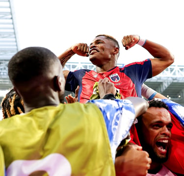 A smiling soccer player in a blue and red jersey celebrates a goal with teammates in a stadium.