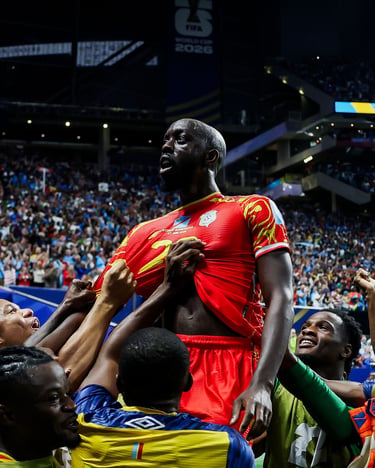 A soccer player in a red jersey celebrates a goal with teammates in a packed stadium.