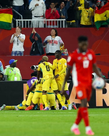 Ghana national football team players celebrating a goal on the field while fans cheer in the stadium stands.