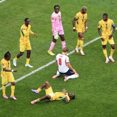 Ghana football players in yellow jerseys stand near Harry Kane on the grass field during a soccer match.