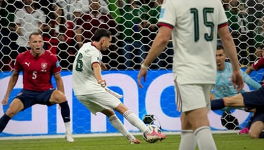 A soccer player in a white jersey takes a penalty kick toward a goalkeeper in a professional match.