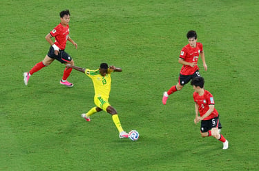 A soccer player in a yellow jersey dribbles past South Korean defenders on a green grass field.
