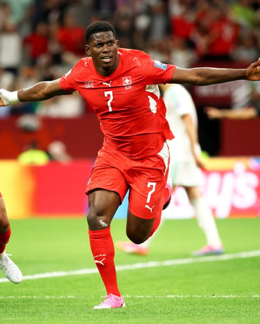 Breel Embolo celebrates a goal for Switzerland in a red soccer jersey on a stadium pitch.
