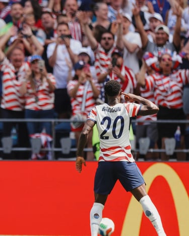 USMNT player Folarin Balogun celebrates a goal in front of cheering soccer fans in the stadium.