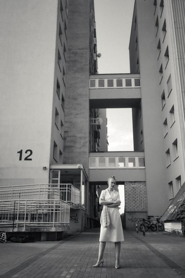 A black and white photo of a woman in a dress standing before a brutalist apartment building.