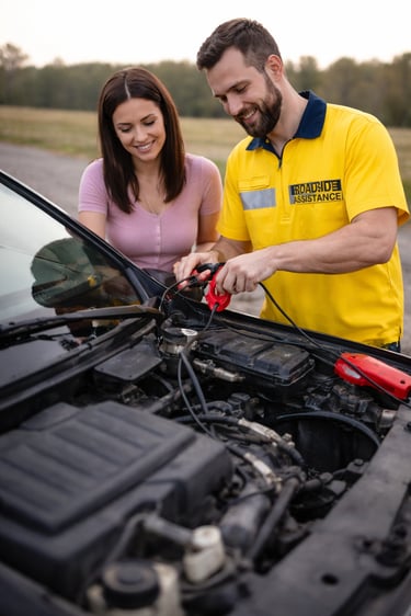 Roadside assistance technician using jumper cables to jump start a smiling woman's car battery.