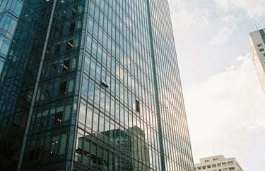 A perspective shot of a sleek glass corporate office building in a major Latin American city under a bright sky. Professional, secure, and modern urban environment. Tones of teal and off-white.