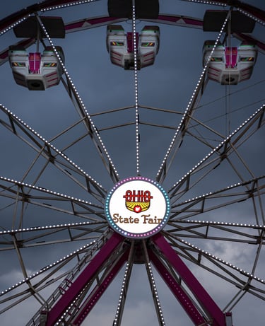 A Ferris wheel at the Ohio State Fair with illuminated passenger gondolas against a cloudy evening sky.