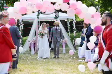 Brautpaar geht nach Trauung unter rosa Ballonbogen hindurch; im Hintergrund ein weißer Pavillon im Garten.