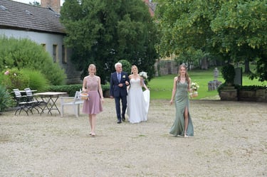 Hochzeitsfotograf Möhnesee: Brautvater führt Braut zur Trauung; vorneweg gehen Blumenmädchen im Garten eines Gutshofs.