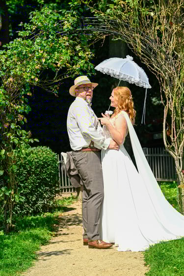 Hochzeitsfotograf Möhnesee: Brautpaar posiert unter grünem Rosenbogen; Braut hält weißen Vintage-Sonnenschirm aus Spitze.
