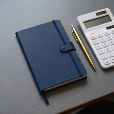 A top-down photographic view of a minimalist workspace in a North American office. A single navy blue notebook, a white calculator, and a high-end gold ballpoint pen are arranged neatly on a charcoal desk surface.