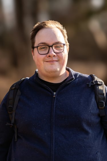 A smiling man with glasses wearing a blue zip-up sweater and a backpack outdoors.