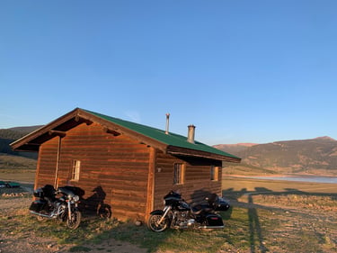 Zwei Harley, die bei Sonnenuntergang vor einer rustikalen Blockhütte in einem Bergtal geparkt sind.