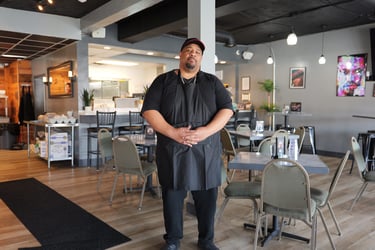 Portrait of a male chef wearing a black apron standing in a modern restaurant dining room.