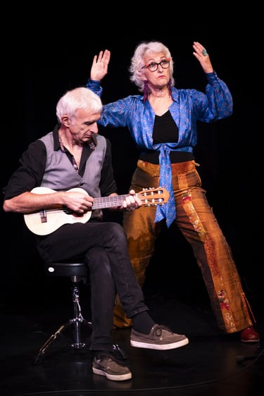 A senior man plays a small guitar while an expressive woman performs a theatrical dance on stage.
