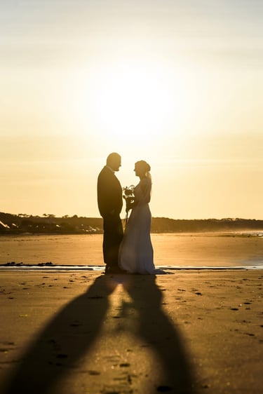 Silhouette of a bride and groom standing on a sandy beach during a golden sunset wedding ceremony.