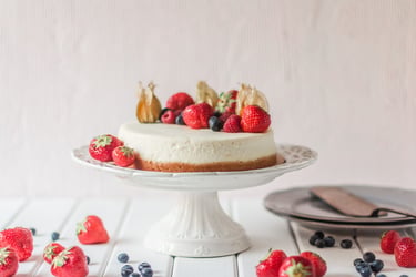 Fresh homemade cheesecake topped with strawberries and blueberries on a white pedestal stand.