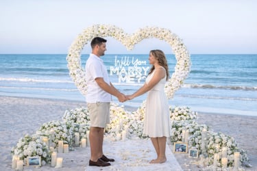 A romantic beach marriage proposal with a white floral heart arch, candles, and neon sign.