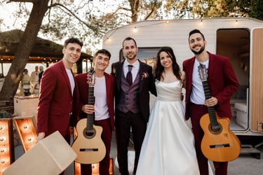 Smiling bride and groom posing with live wedding band guitarists in red suits outdoors.
