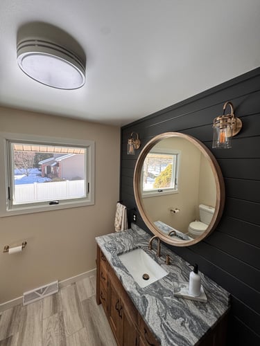 Modern bathroom vanity with a granite countertop, round wood mirror, and black shiplap accent wall.