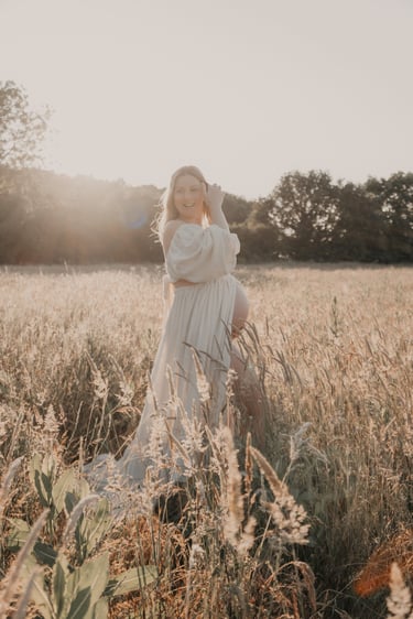 Smiling pregnant woman in a white dress posing in a golden field during a sunset maternity photoshoot.