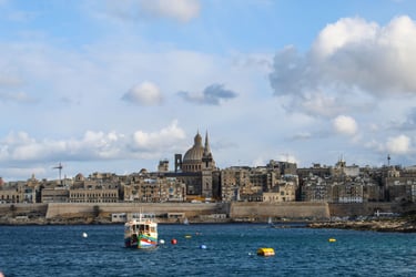 A traditional Maltese boat floats in Valletta harbor with the Basilica of Our Lady of Mount Carmel in the background.