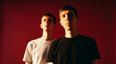 Two men posing in t-shirts against a red background with dramatic warm lighting.