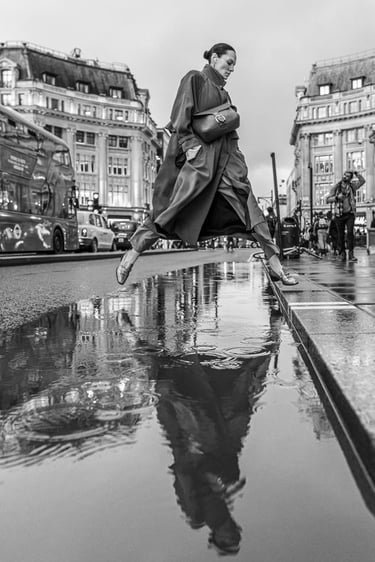 Black and white candid street scene capturing lady leaping to the kerb over a puddle.