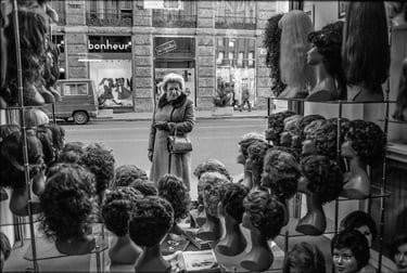 Candid black and white photo of older lady admiring wigs in a shop window.