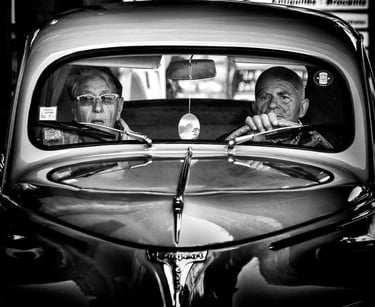 Street photography in black and white, capturing an older couple through the windscreen of a car.
