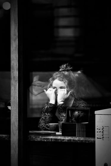 Monochrome image of a lone girl in a coffee shop window wiping tears from her eyes.