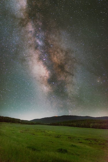 Milky Way galaxy stretching across a starry night sky above a dark forest and green meadow.