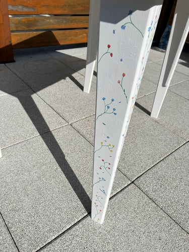 Hand-painted white table leg featuring delicate colorful floral designs on a sunny stone patio.