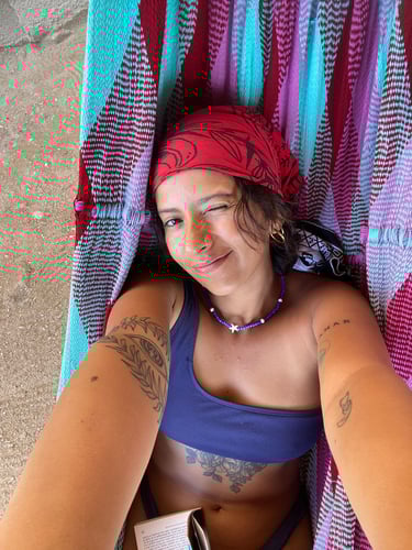 Mujer tatuada con un pañuelo rojo guiñando el ojo mientras se relaja en una colorida hamaca afuera.