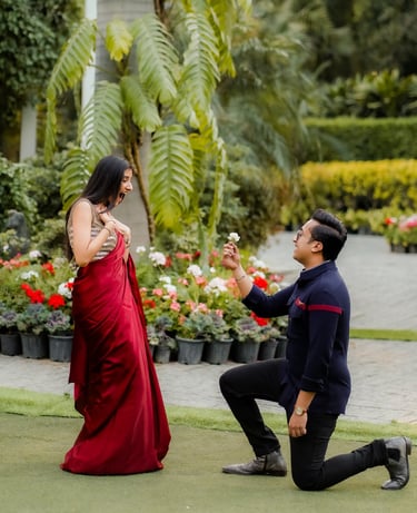 A man on one knee proposing to a woman in a red saree in a lush tropical garden.