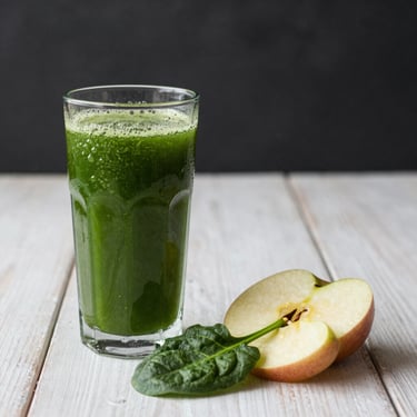 A refreshing green detox juice in a tall glass next to a handful of fresh spinach and apple slices on a white wooden table.