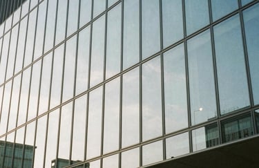 A sleek, modern glass building facade reflecting a clear sky, showing geometric lines and a sense of architectural strength and transparency. Light grayish blue and white tones. Global / Spanish-speaking.