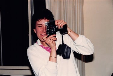A woman in a white sweater smiling while taking a photo with a vintage 35mm film camera.