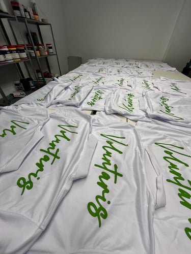 Custom screen printed white volunteer t-shirts with green cursive text laid out in a workshop.