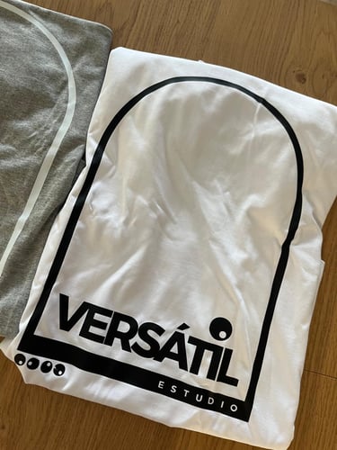 Folded Versátil Estudio white cotton t-shirt with black arch logo on a wooden surface.