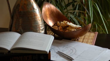 Astrology book and birth chart diagrams next to a copper bowl of potpourri on a desk.