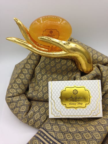 Luxury Achara soap bar held by a gold hand sculpture on a Thai silk fabric background.