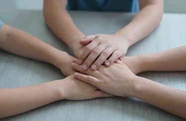 A close-up shot of a North American / US family's hands joined together over a kitchen table, symbolizing protection and unity. The scene features soft gray-blue lighting.