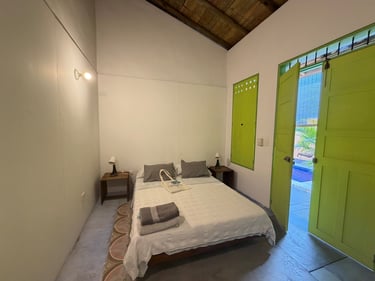Rustic tropical bedroom with white bedding, wooden ceiling, and bright green shutters and door.
