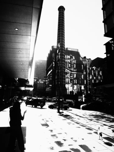 Cityscape with dramatic shadows. A tall chimney rises among buildings. A silhouetted person and a bird add to the scene.