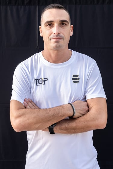 A smiling male fitness coach in a white polo shirt posing with crossed arms against a black background.